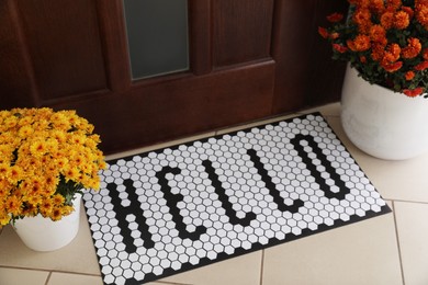 Stylish door mat with word HELLO and beautiful flowers on floor