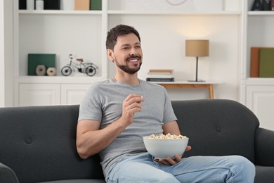 Photo of Happy man with bowl of popcorn watching movie via TV on sofa at home