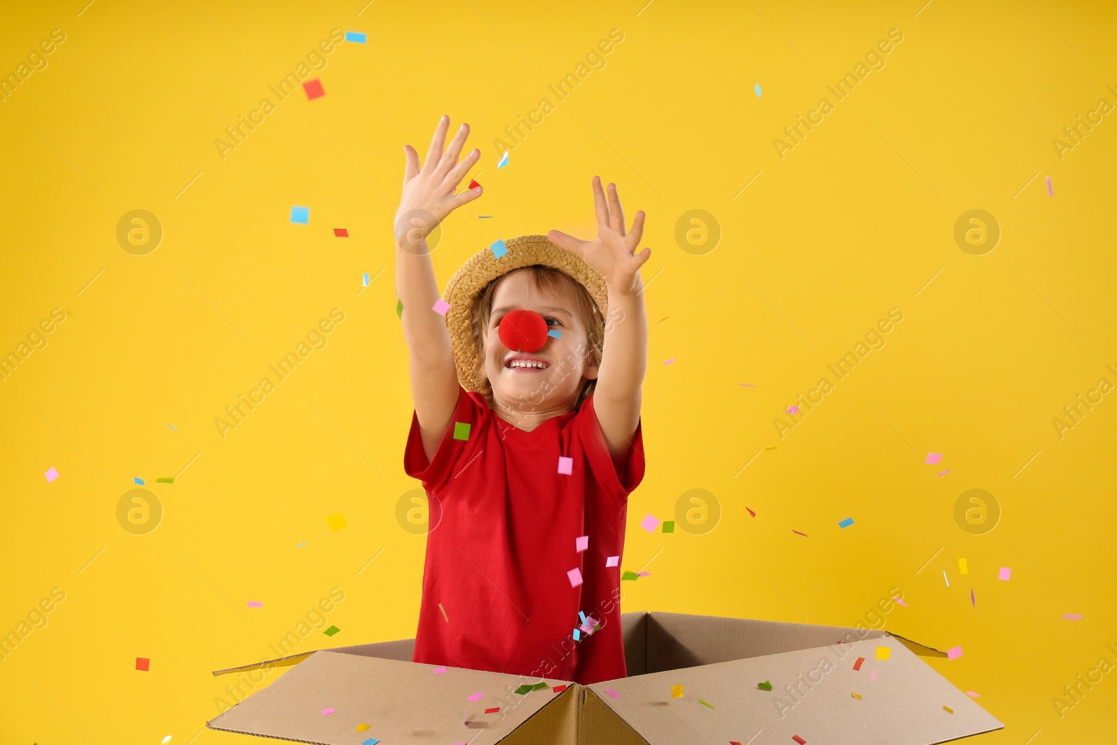 Photo of Little boy with clown nose in cardboard box under confetti shower on yellow background. April fool's day