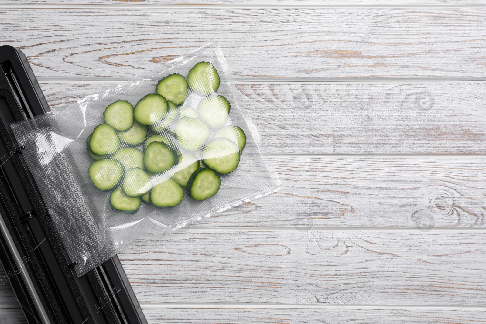 Photo of Vacuum packing sealer with plastic bag of cucumbers on white wooden table, flat lay. Space for text