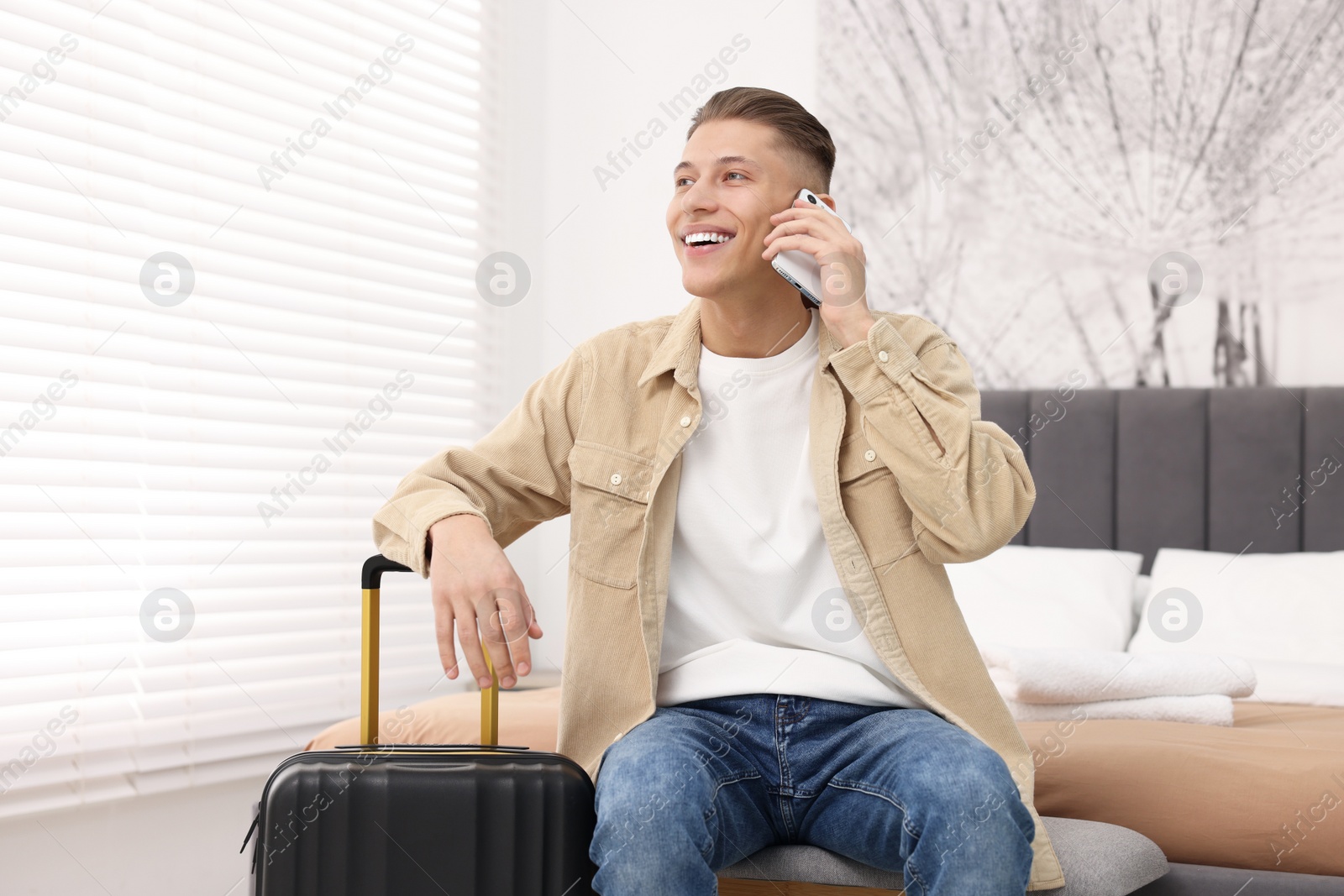 Photo of Smiling guest with suitcase talking on smartphone in stylish hotel room