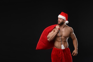 Muscular young man in Santa hat holding bag with presents on black background, space for text