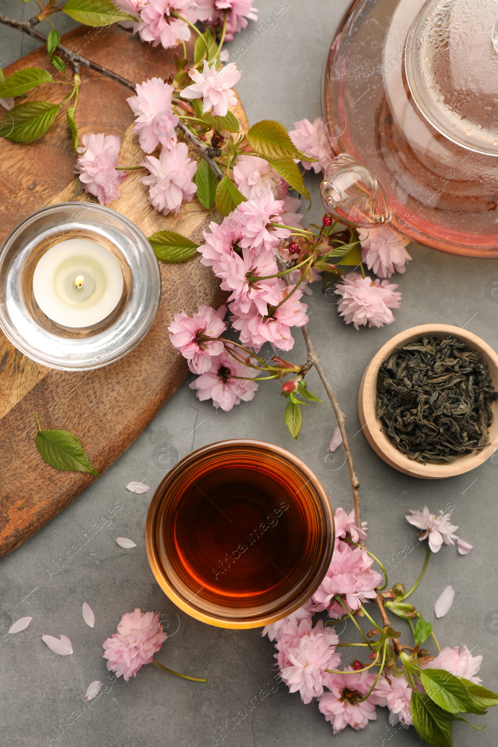 Photo of Traditional ceremony. Cup of brewed tea, burning candle, teapot and sakura flowers on grey table, flat lay