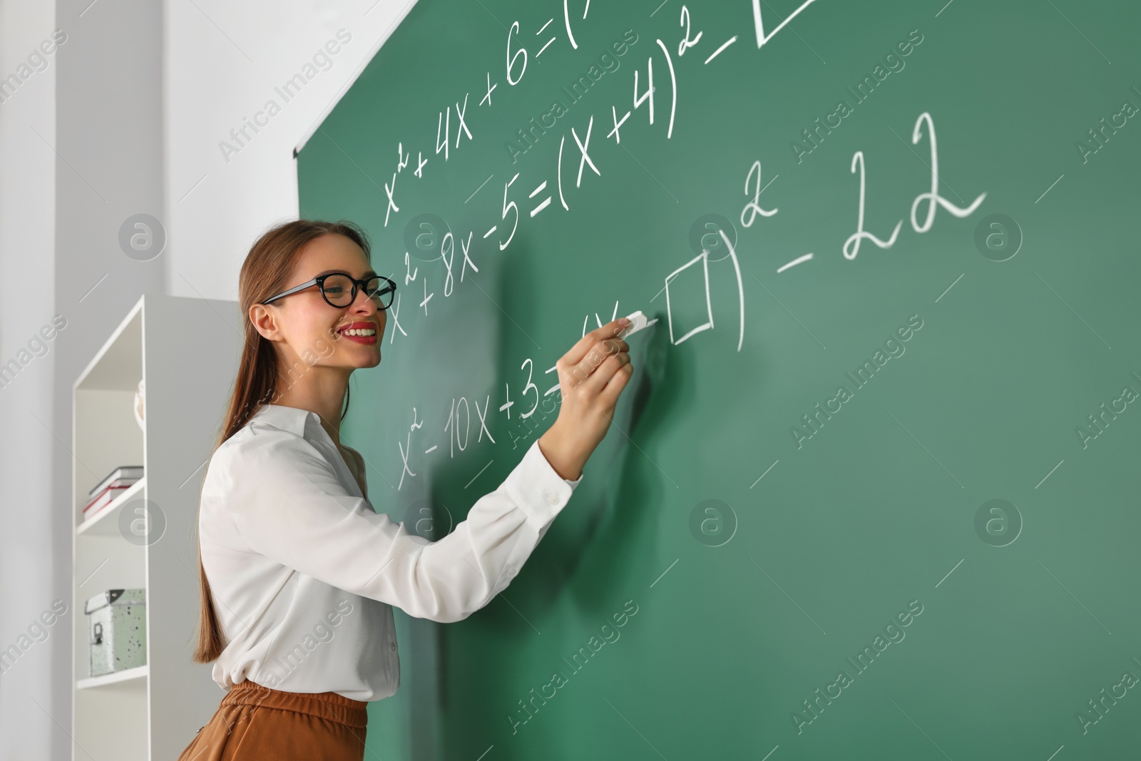 Photo of Young math’s teacher writing mathematical equations near chalkboard in classroom