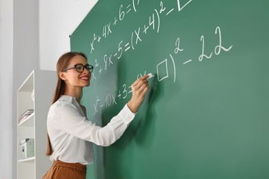 Photo of Young math’s teacher writing mathematical equations near chalkboard in classroom