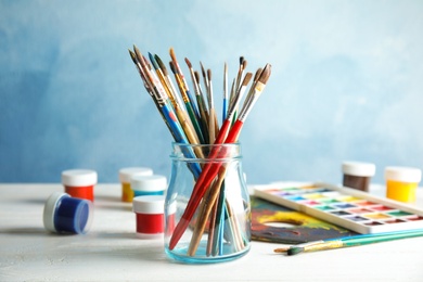 Glass jar with brushes and paints on table against color background