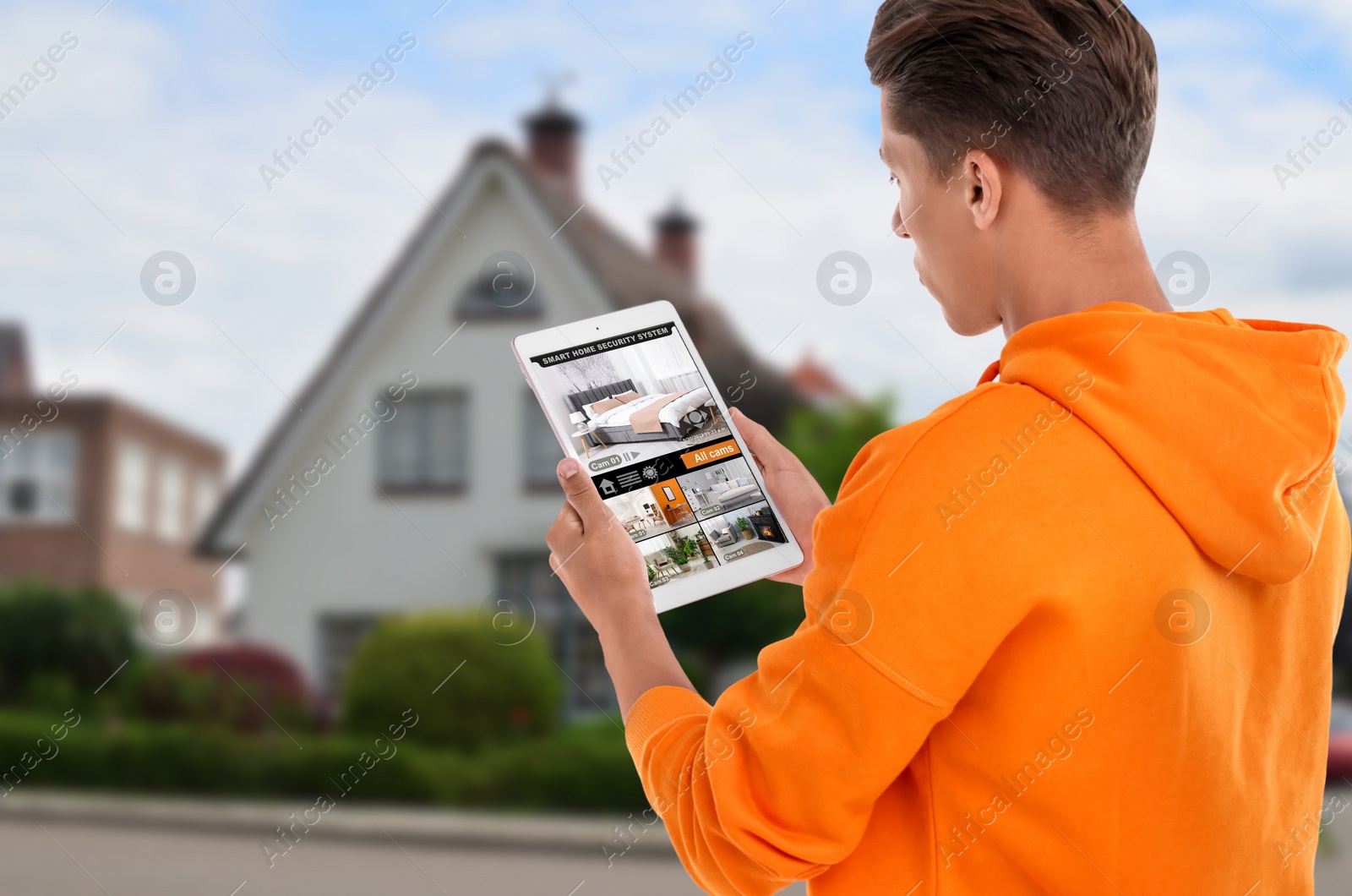Image of Man using smart home security system on tablet computer near house outdoors. Device showing different rooms through cameras