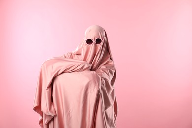 Photo of Glamorous ghost. Woman in sheet with sunglasses on pink background, space for text