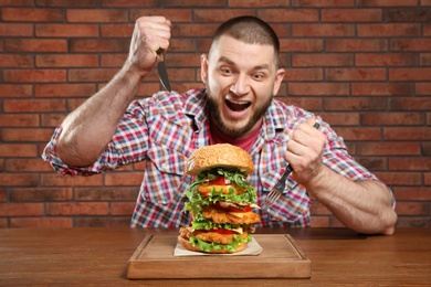 Photo of Young hungry man with cutlery and tasty huge burger at table