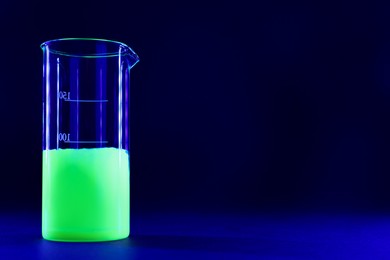 Photo of Laboratory beaker with luminous liquid on dark blue background, space for text