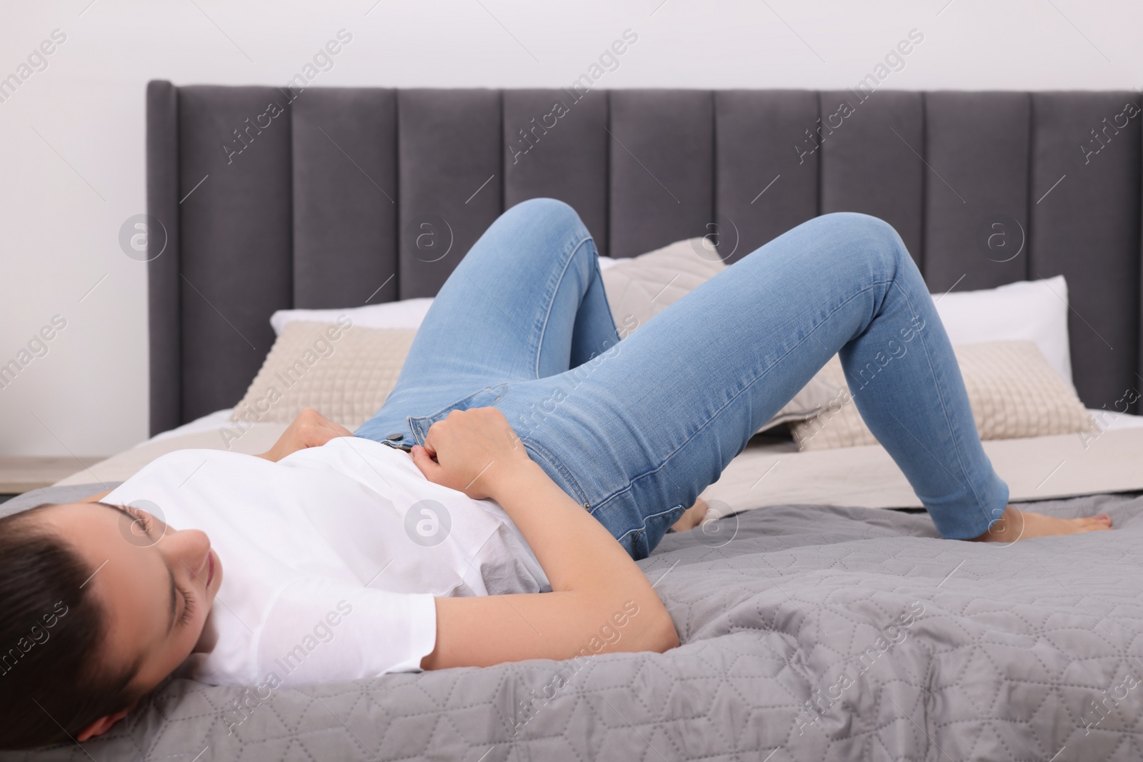 Photo of Woman trying to put on tight jeans on bed at home