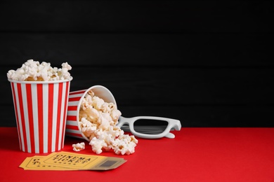 Photo of Cups with delicious popcorn, eyeglasses and tickets on red table against black background
