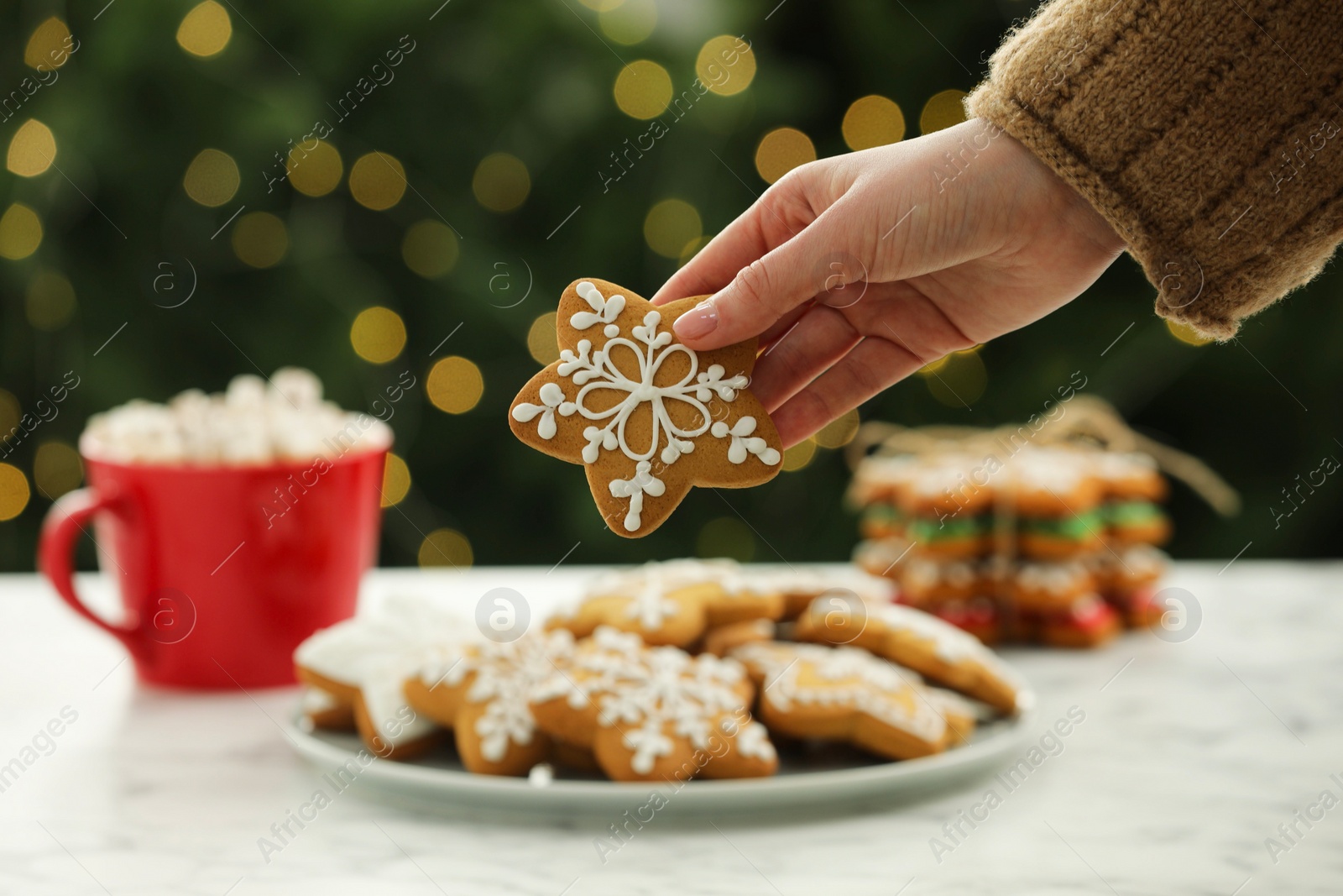 Photo of Woman with decorated cookie at table against blurred Christmas lights, closeup