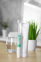 Electric toothbrush, tube with paste and glass of water on wooden table in bathroom