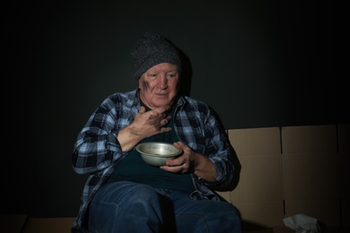 Photo of Poor senior man with bowl on floor near dark wall