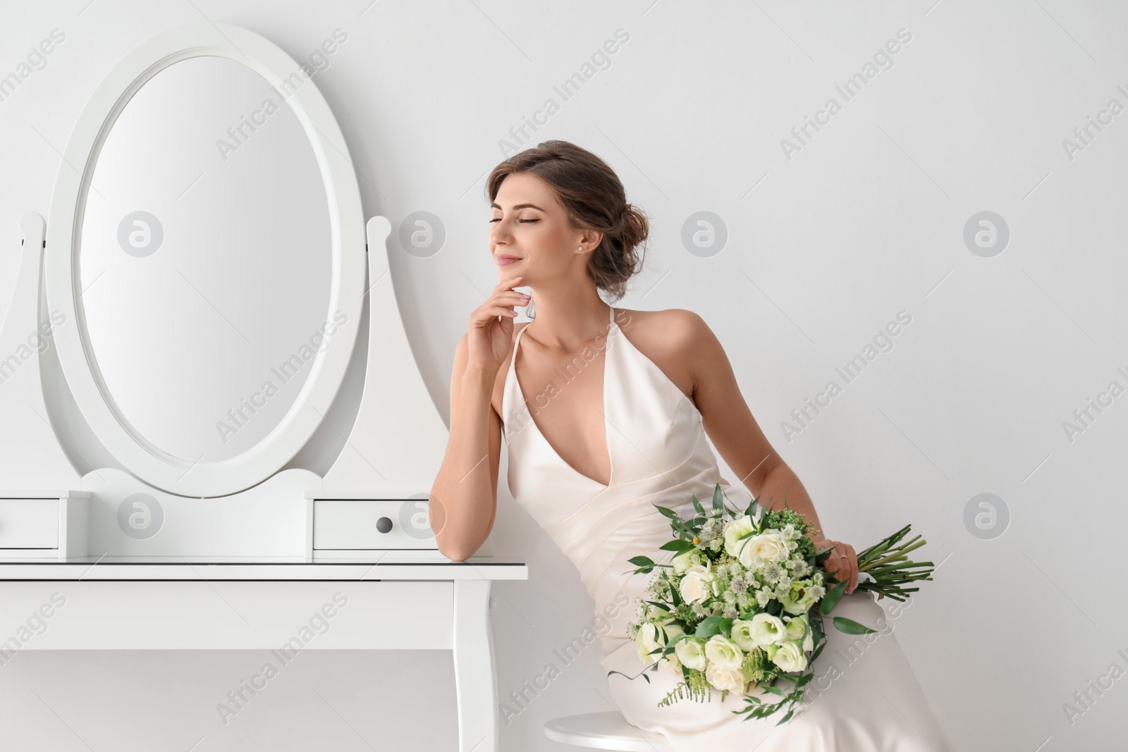 Photo of Young bride in wedding dress with beautiful bouquet near mirror indoors