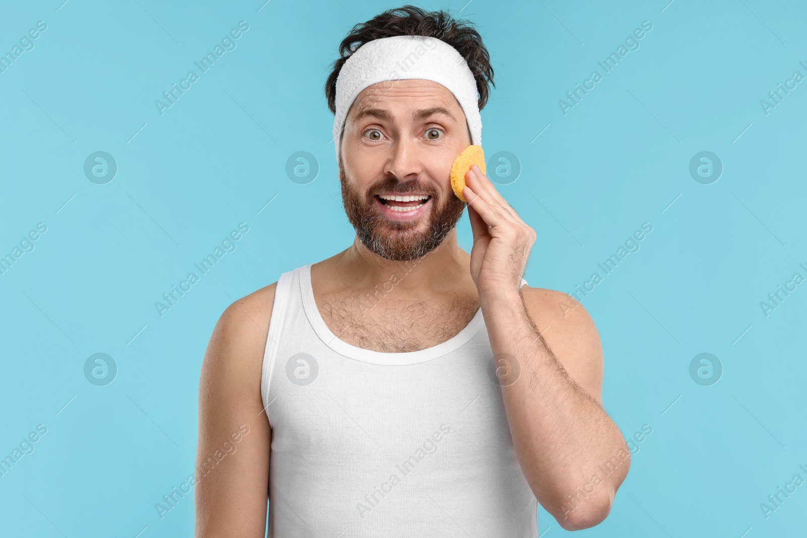 Photo of Man with headband washing his face using sponge on light blue background