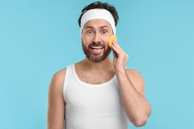 Man with headband washing his face using sponge on light blue background