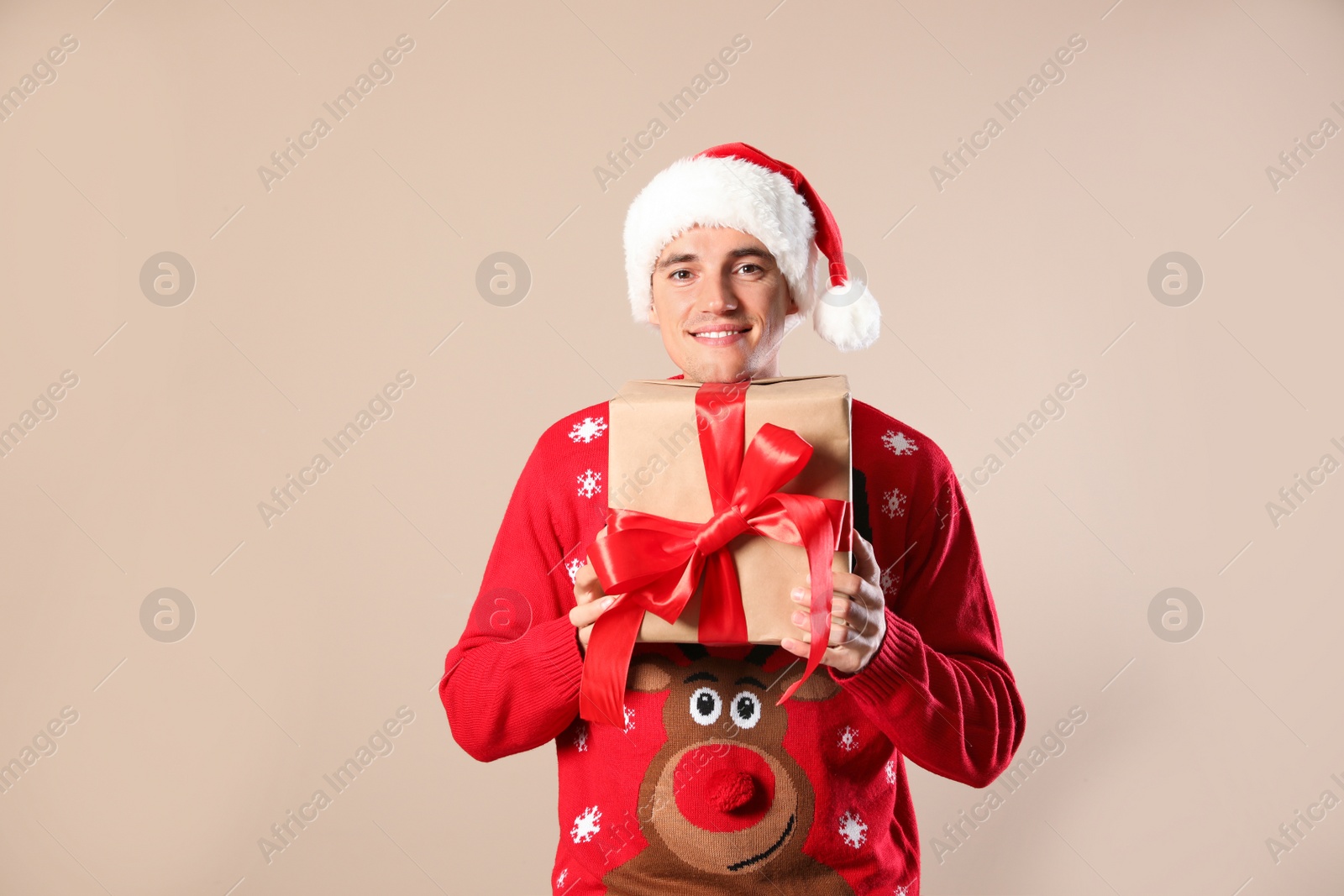 Photo of Happy man in Christmas sweater and Santa hat holding gift box on beige background