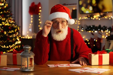 Emotional Santa Claus at his workplace. Letters and gift boxes on table in room decorated for Christmas