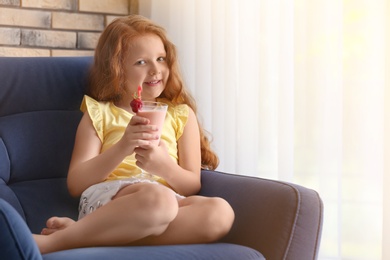 Photo of Little girl with glass of delicious milk shake in armchair indoors