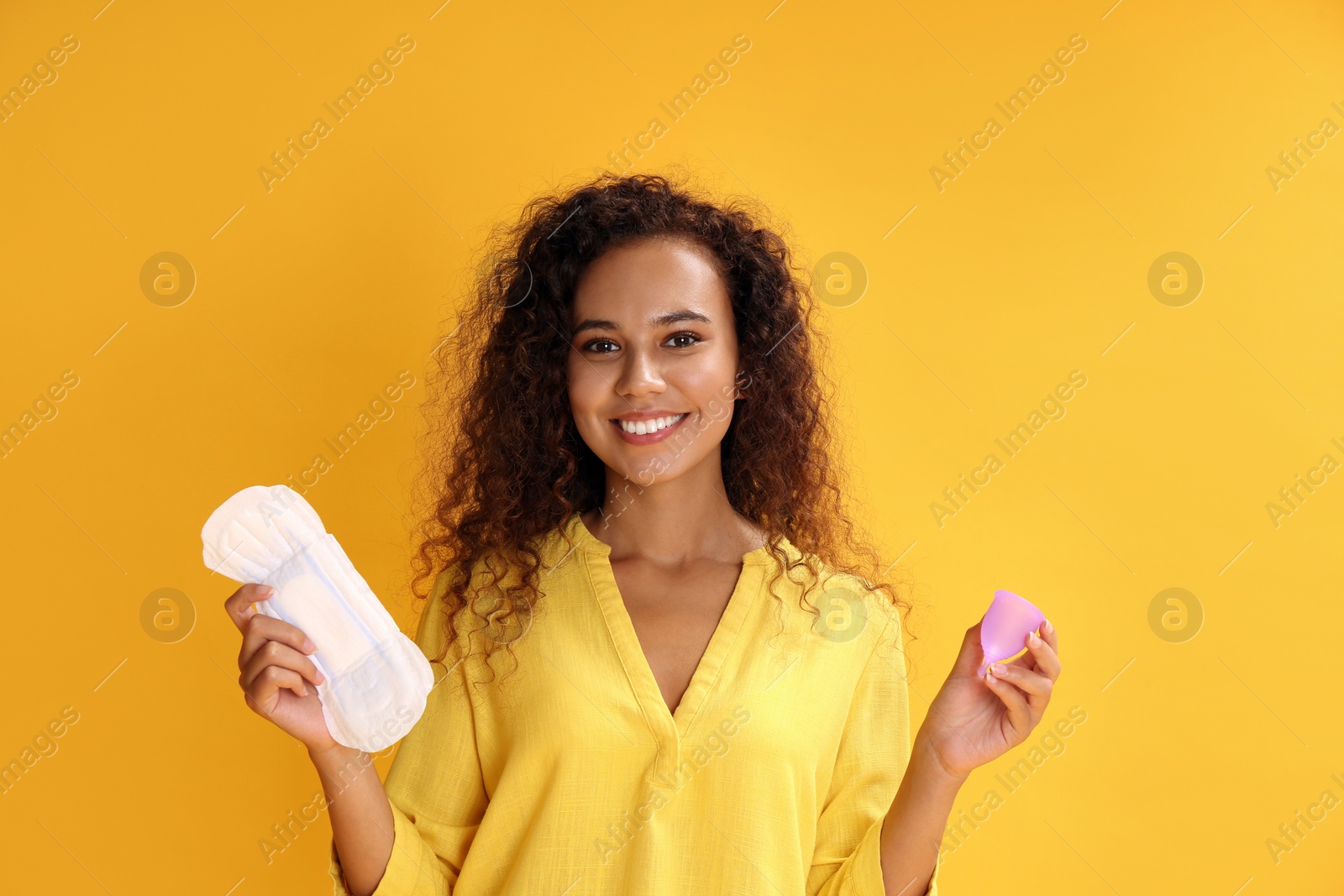 Photo of Young African American woman with menstrual cup and pad on yellow background