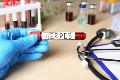 Doctor in glove holding test tube with word Herpes at wooden table, closeup