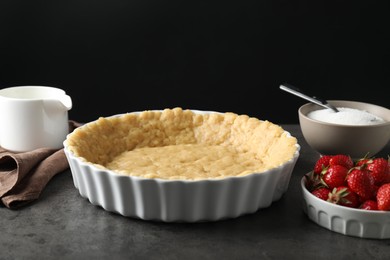 Photo of Making shortcrust pastry. Raw dough in baking dish, strawberries, sugar and pitcher on grey table