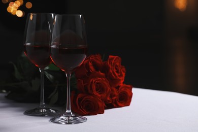 Photo of Glasses of red wine and rose flowers on white table against blurred lights, space for text. Romantic atmosphere