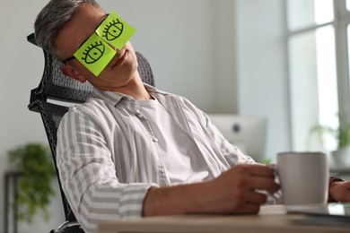 Photo of Man with fake eyes painted on sticky notes snoozing at workplace in office