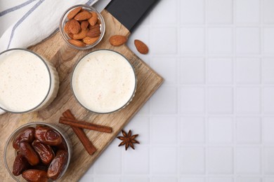 Glasses of delicious date smoothie, dried fruits, spices and nuts on white tiled table, flat lay. Space for text