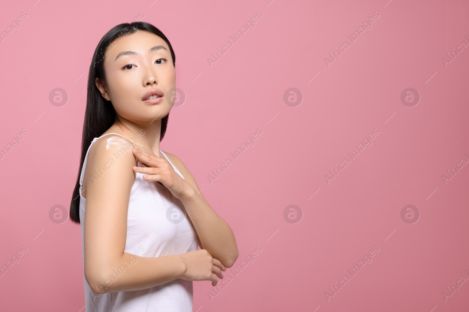 Photo of Beautiful young Asian woman applying body cream onto shoulder on pink background, space for text