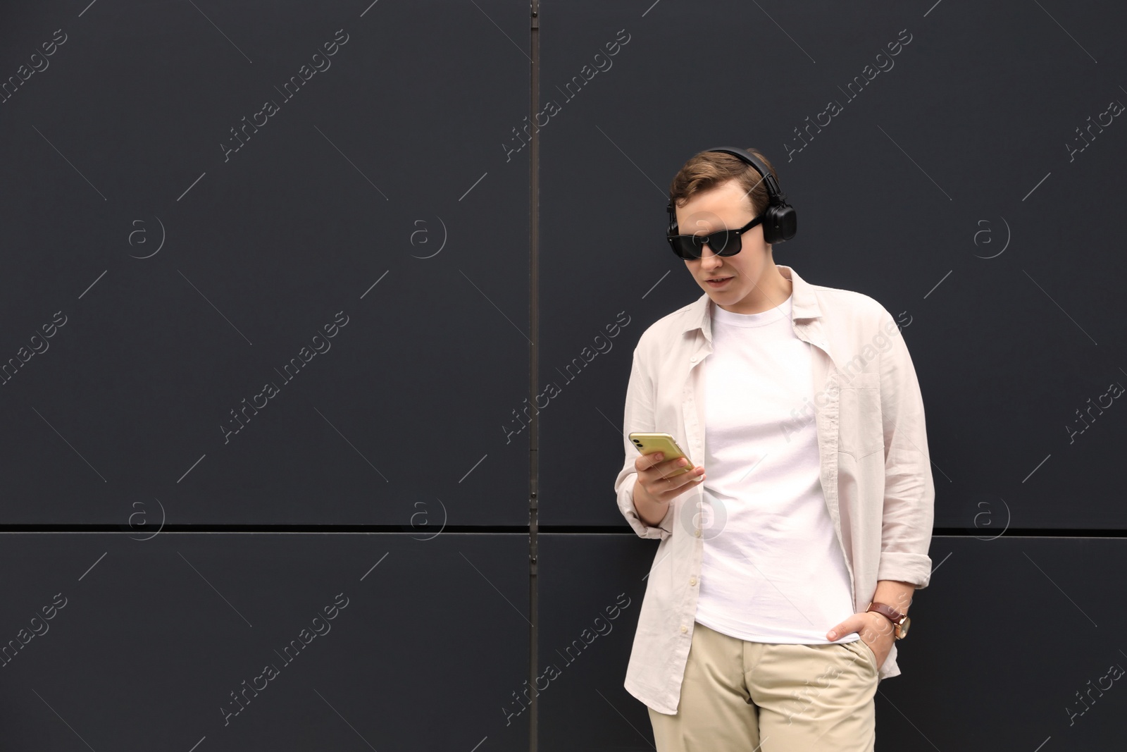 Photo of Handsome young man with headphones and modern smartphone near black wall. Space for text