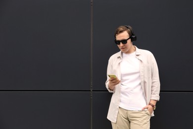 Handsome young man with headphones and modern smartphone near black wall. Space for text