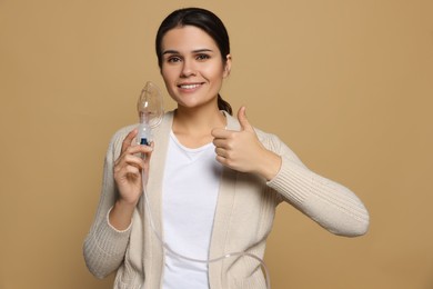Photo of Happy young woman with nebulizer showing thumb up on dark beige background