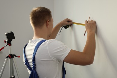 Photo of Worker using cross line laser level, pencil and tape for accurate measurement on light wall, back view