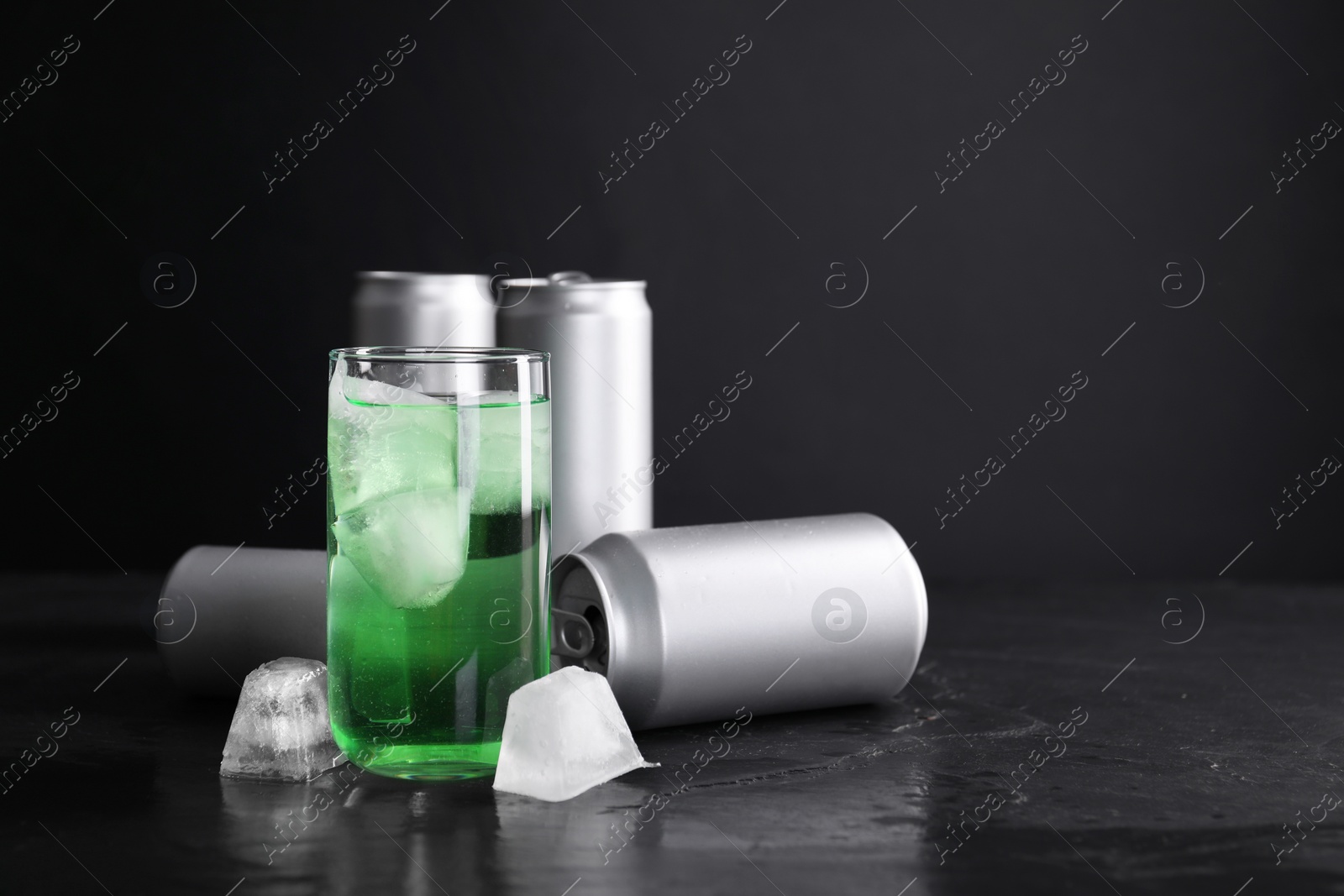 Photo of Tasty energy drink with ice cubes in glass and aluminium cans on black table. Space for text