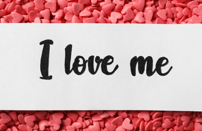 Piece of paper with handwritten phrase I Love Me on pink heart shaped sprinkles, top view