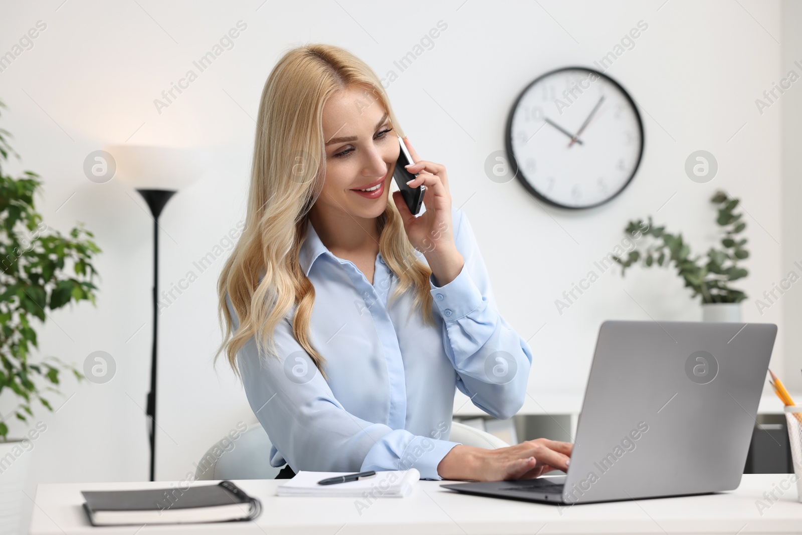 Photo of Happy secretary talking on smartphone while working with laptop in office