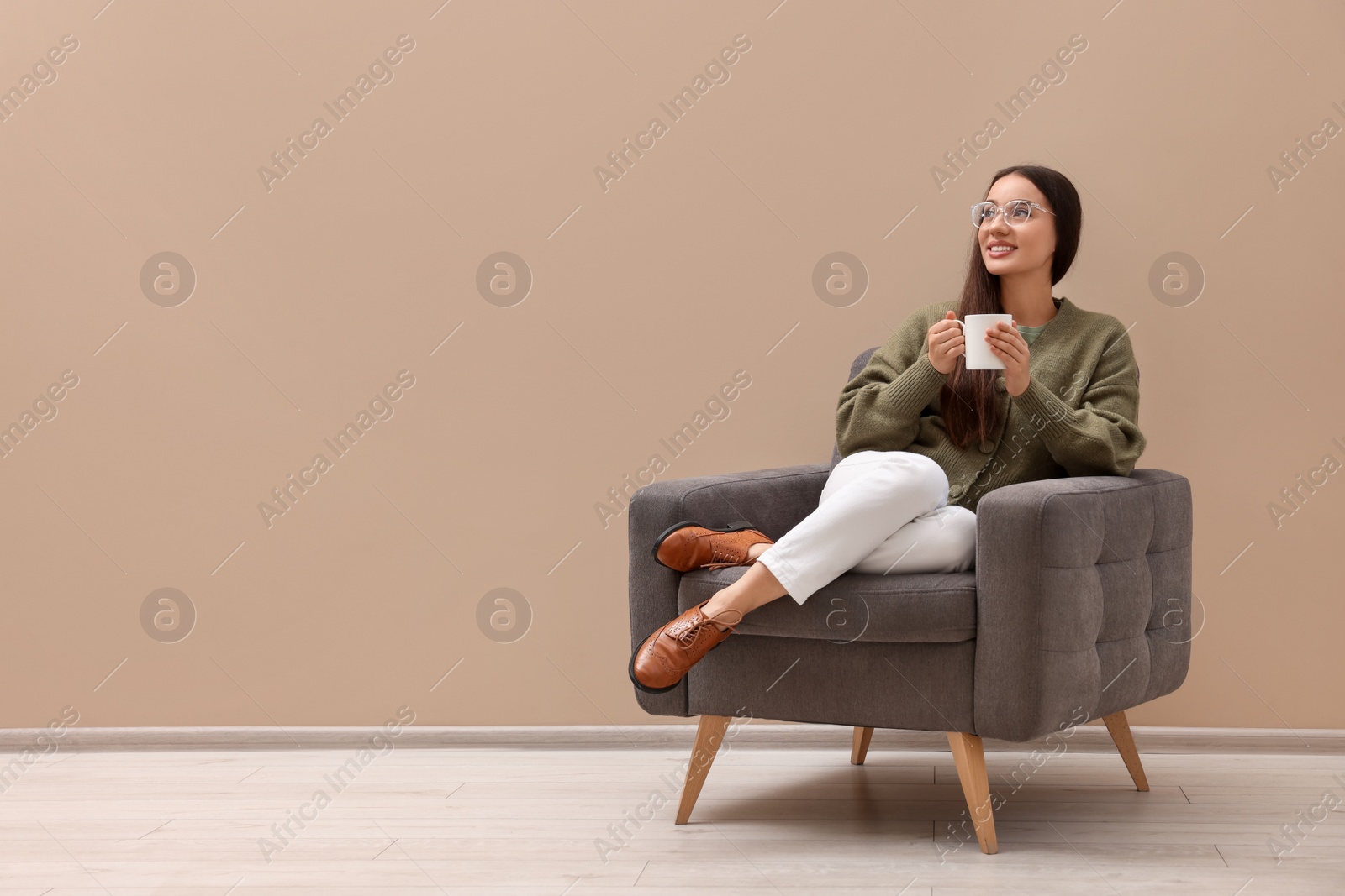 Photo of Beautiful woman with cup of drink sitting in armchair near beige wall indoors, space for text