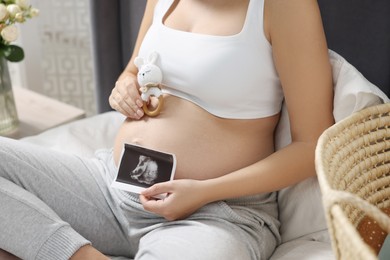Photo of Pregnant woman with ultrasound picture of baby and bunny toy sitting on bed indoors, closeup