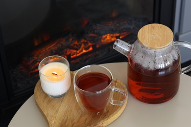 Photo of Teapot, cup of aromatic tea and burning candle on white table near fireplace indoors