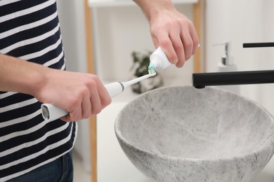 Man squeezing toothpaste from tube onto electric toothbrush above sink in bathroom, closeup