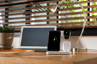Mobile phone, earphones and smartwatch charging with wireless pad on wooden desk, space for text. Modern workplace device