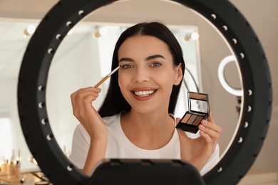 Beautiful young woman applying eye shadow with brush indoors, view through ring lamp