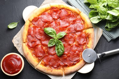 Photo of Tasty pepperoni pizza, sauce, basil and cutter on black textured table, flat lay