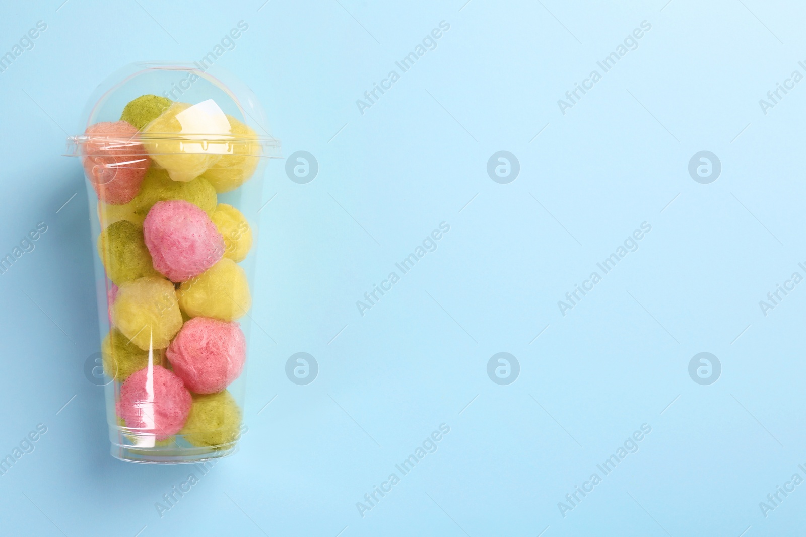 Photo of Plastic cup with color cotton balls on light blue background, top view and space for text. Sweet candy