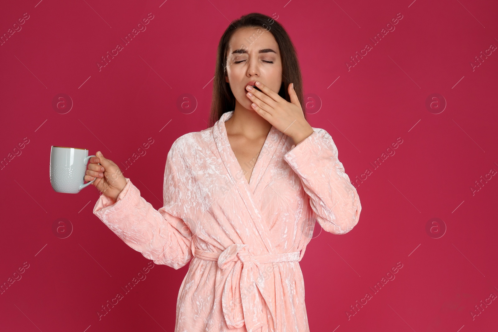 Photo of Young woman in bathrobe with cup of coffee yawning on crimson background