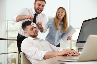 Photo of Coworkers sticking note with word Fool to colleague's face in office. Funny joke