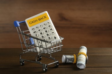 Photo of Rolled banknotes and calculator with sign Cash Back in small shopping cart on wooden table. Space for text